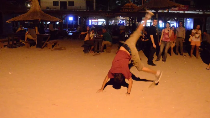 Young man doing a cartwheel on the sand at night, with a small crowd watching near beach bars.