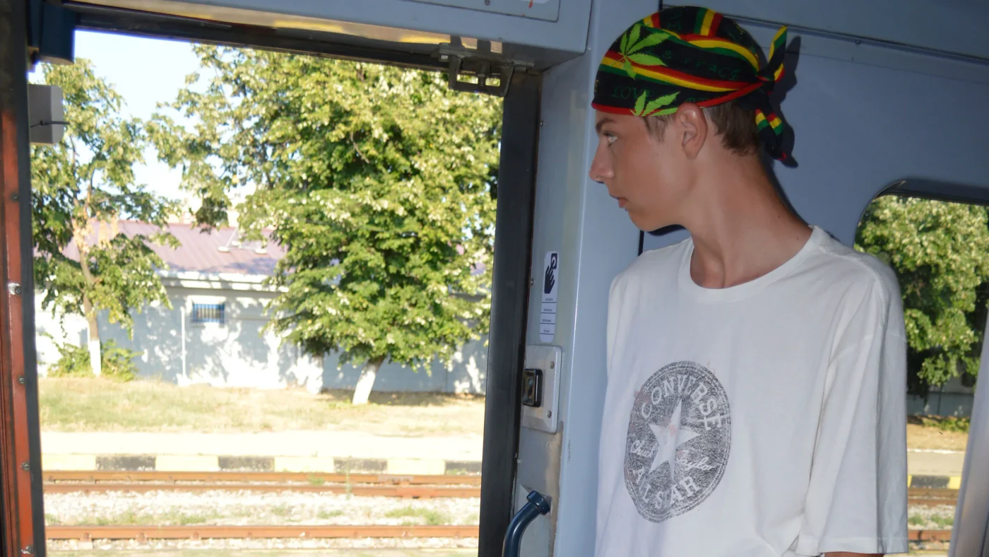 Teen standing in the open doorway of a summer train, wearing a colorful reggae bandana and white T-shirt, looking out at the station tracks and trees while traveling to Vama Veche.