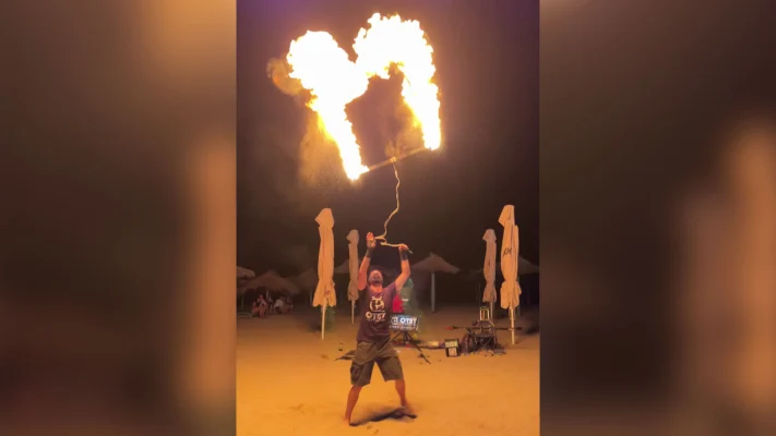 A fire juggler balances a flaming square frame above his head at night on the beach.