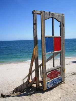 A wooden door with painted panels and a fish symbol standing on the sand, marking the entrance to Cherhana restaurant in Vama Veche.