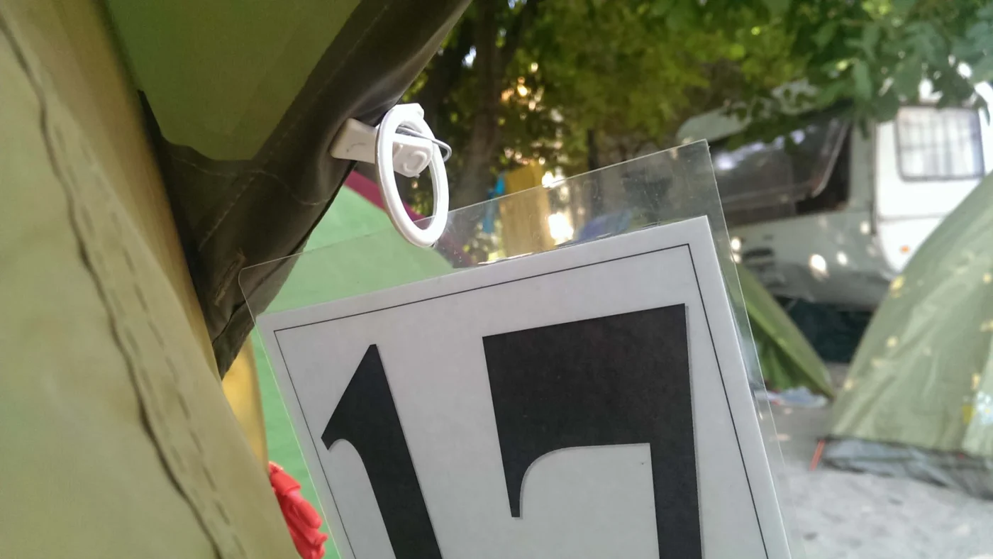 Close-up of a laminated campsite tag “17” clipped to a tent doorway under trees, other tents blurred in the background.