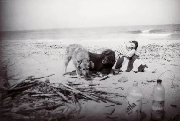 Two young people resting on the beach with a dog, beside driftwood and empty soda bottles, capturing the raw, unfiltered atmosphere of Vama Veche in the 90s.