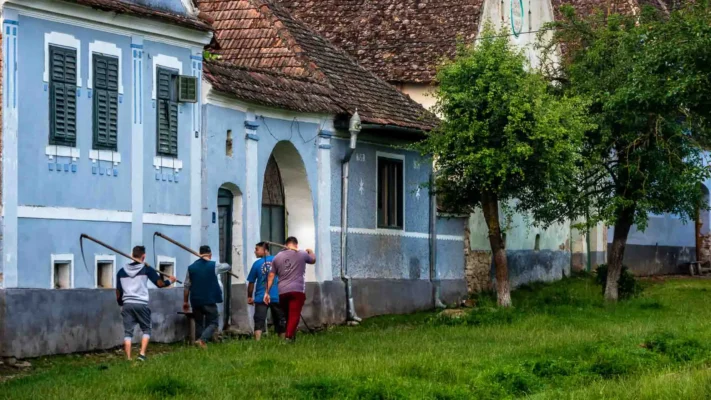 Local residents carrying scythes along the grassy edge of Viscri's main street beside traditional village houses.