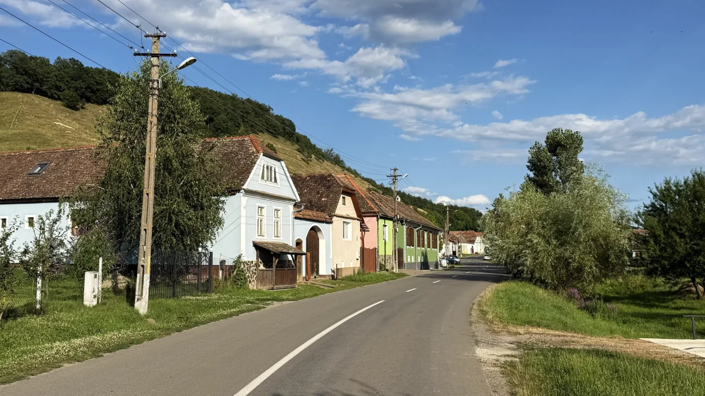 Main road through Cund, Transylvania, lined with colorful traditional houses beneath wooded hills and a summer sky.