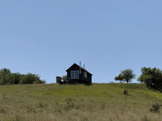 Small wooden cabin standing alone on a grassy hill above Cund, beneath a clear summer sky.