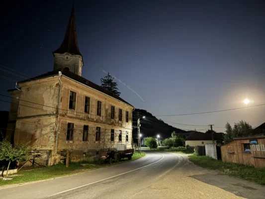 Abandoned church and old village building in Cund illuminated at night beneath the moon and a starry sky.